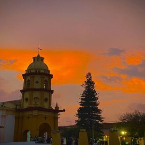 Atardecer sobre la Catedral de San Francisco, con nubes naranjas y azuladas enmarcando la escena. Un pino y siluetas de personas completan el paisaje.
