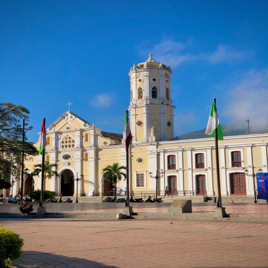 Vista de la Catedral de Santa Ana en Ocaña, Norte de Santander, con su torre y fachada neoclásica, rodeada de banderas y un cielo azul despejado.